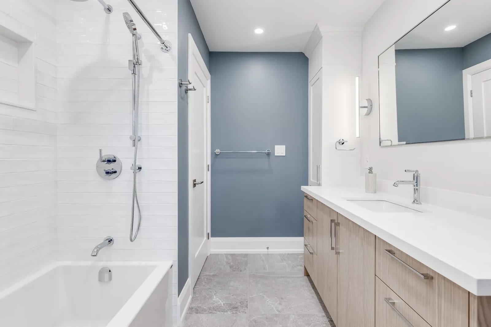 Tub and shower in white stacked tile with a rainfall head and hand shower, the slate blue wall beside it and the light oak vanity opposite.