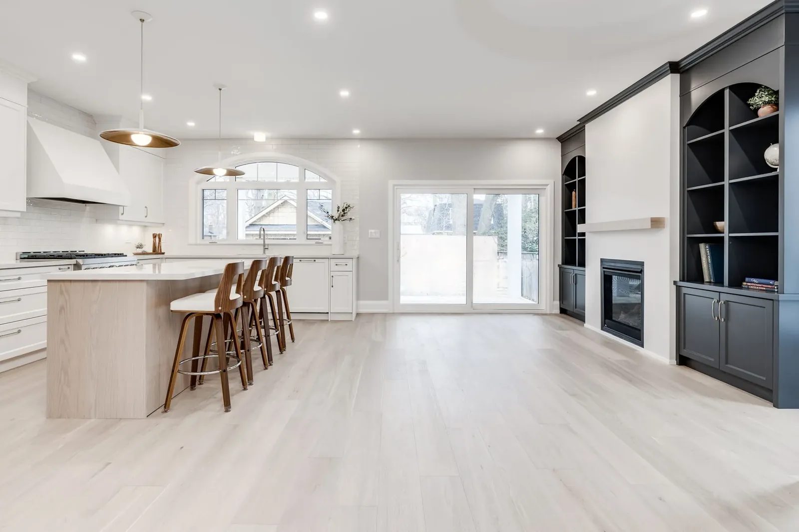 Open-plan main floor with the kitchen island and stools on one side and the charcoal built-ins and fireplace on the other, joined by pale oak floors.