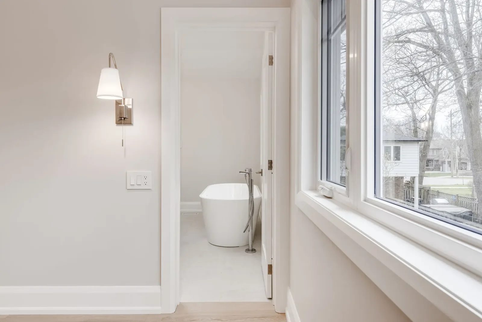 Primary bedroom wall with a polished nickel reading sconce beside a wide window onto the street, the door standing open through to the ensuite and its freestanding tub.