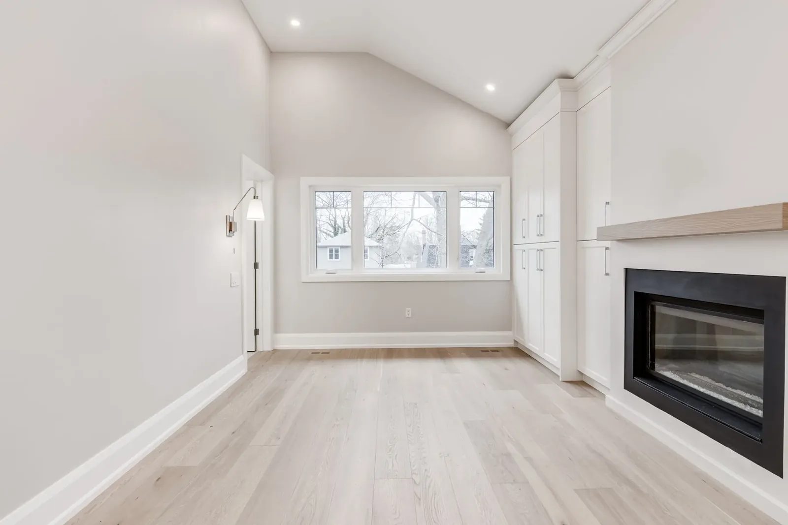 Vaulted primary bedroom with the wall of white built-in cabinetry and the fireplace under an oak shelf, beside a wide window.