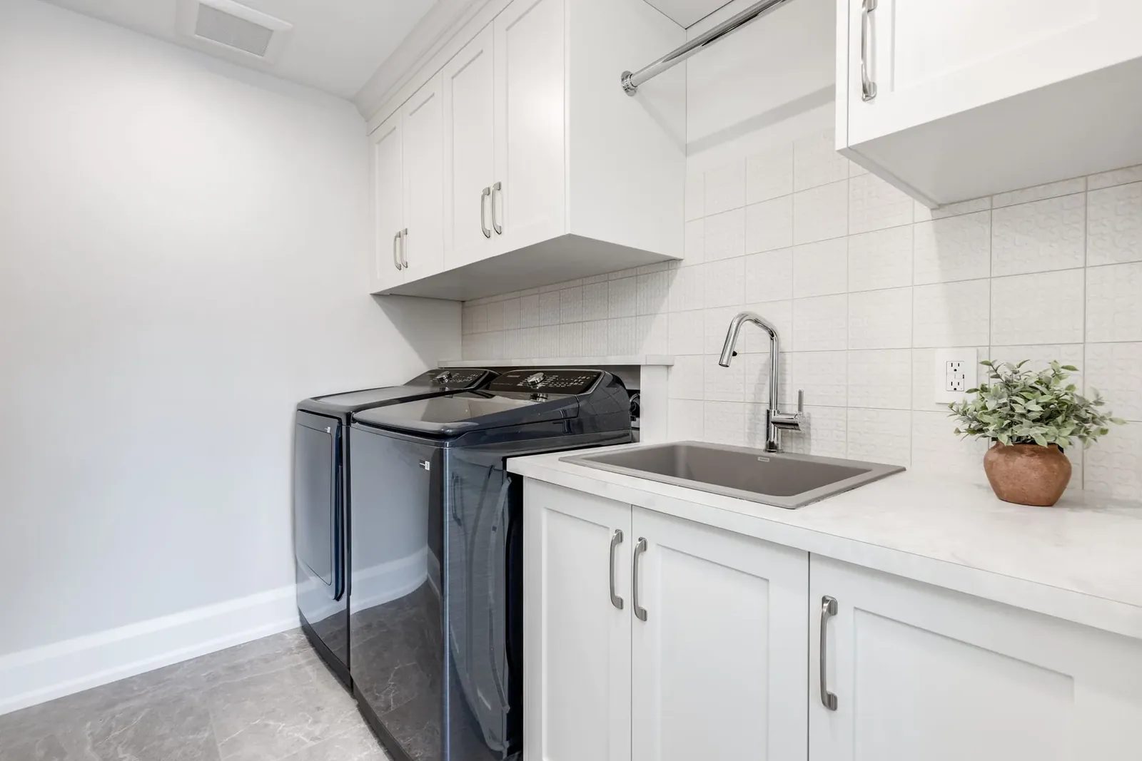 Laundry room with white shaker cabinets over the machines, a sink set into the counter and a textured white tile backsplash.