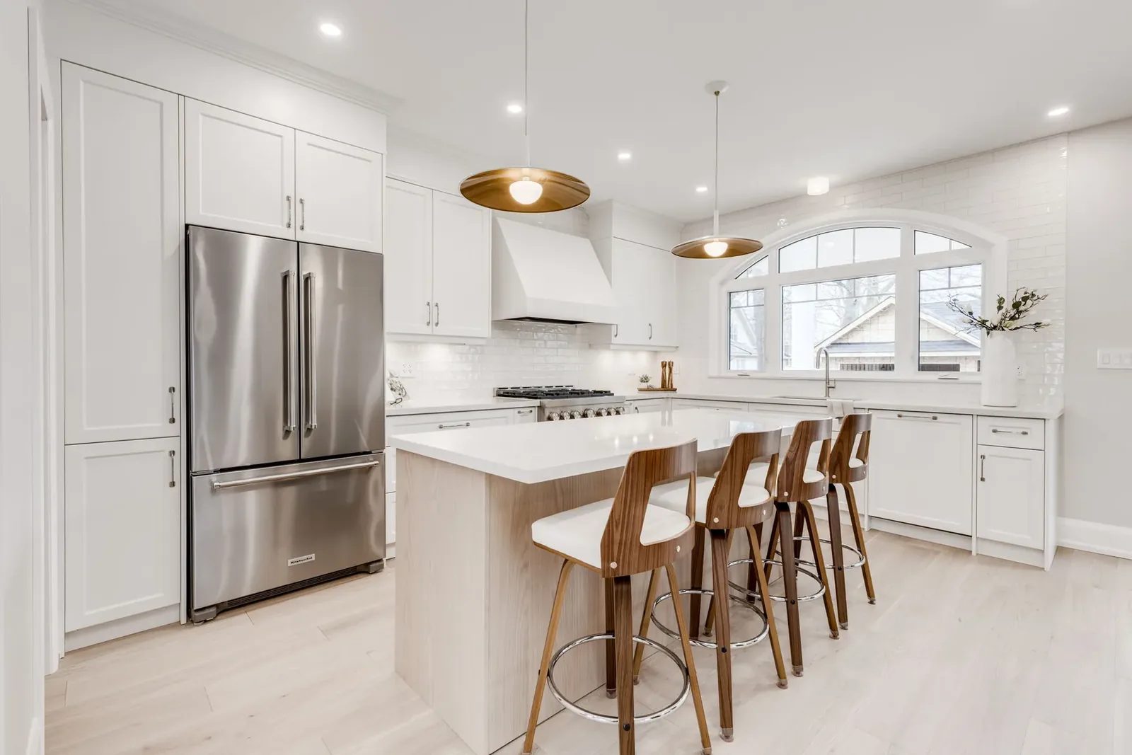 White shaker kitchen with the stainless refrigerator set into tall cabinetry, the oak island and four walnut and white stools under brass saucer pendants, and the arched window over the sink.