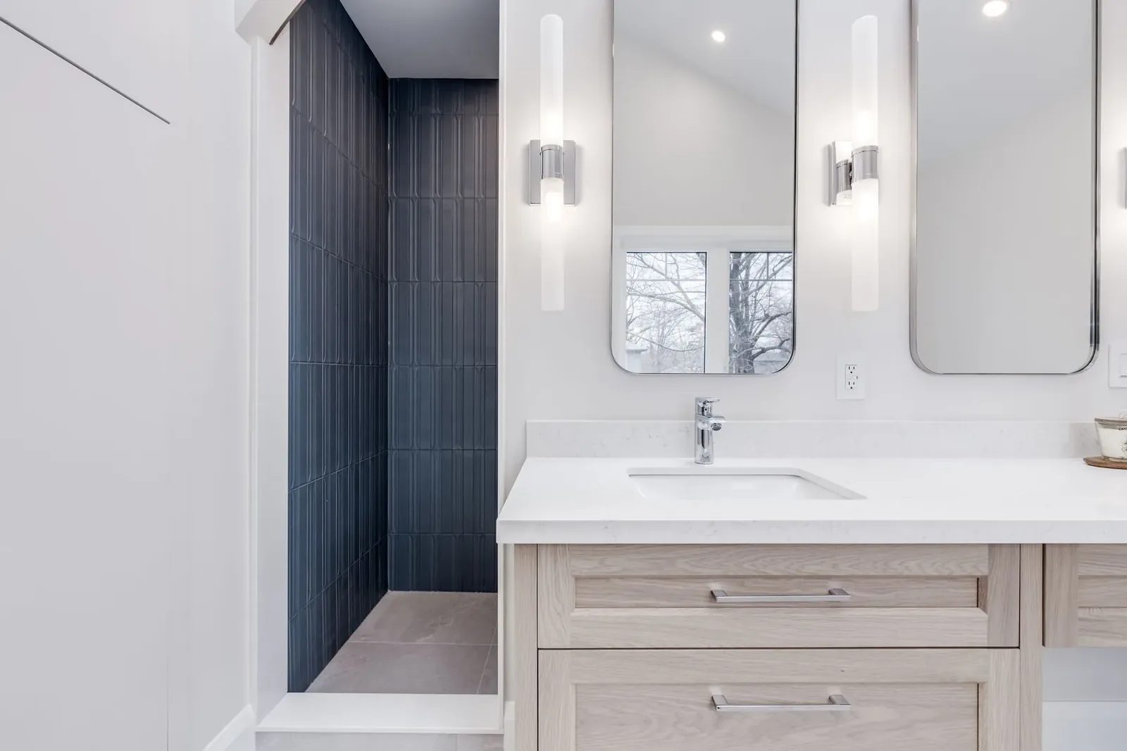 Ensuite vanity in light oak under a quartz counter, twin rounded chrome-framed mirrors between cylinder sconces, and the dark fluted tile shower open alongside.