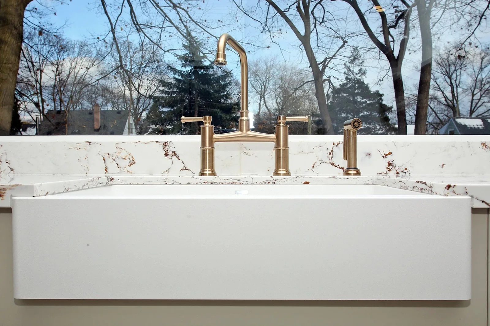 Warm white apron-front sink with brushed brass tap set against veined quartz beneath a broad window.