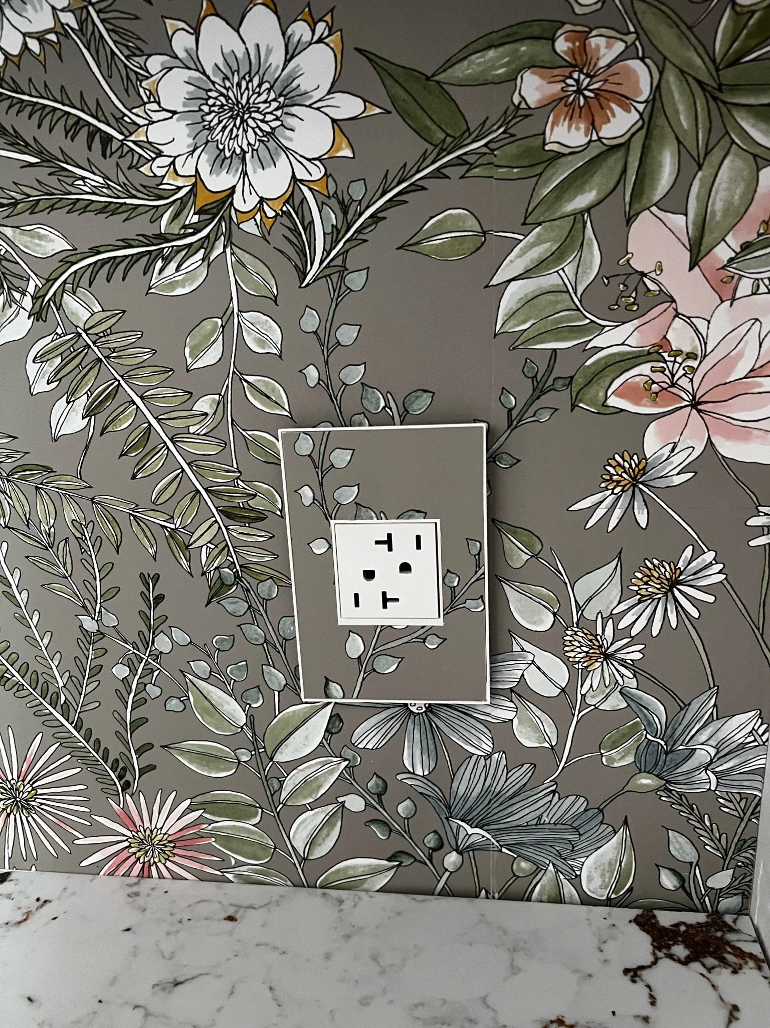 Botanical wallpaper surrounds a GFCI outlet above a white stone counter in a finished bathroom detail.