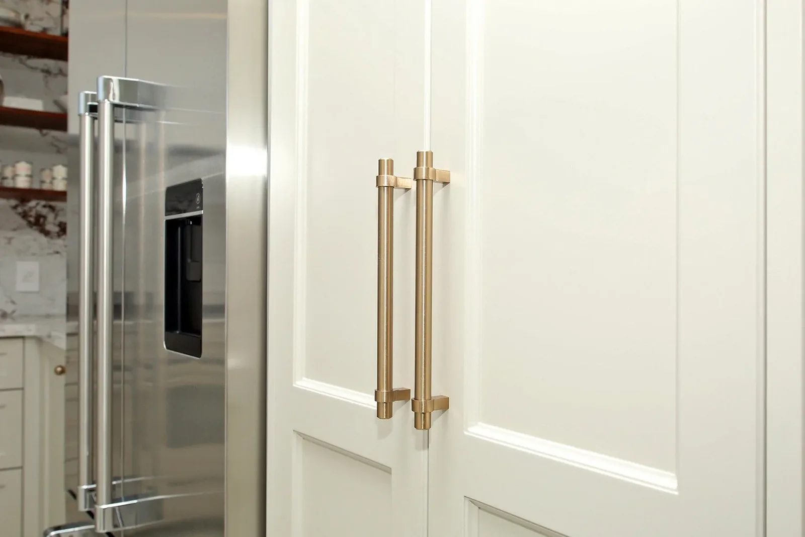 Warm white kitchen cabinet doors with brass pulls sit beside a stainless steel refrigerator.