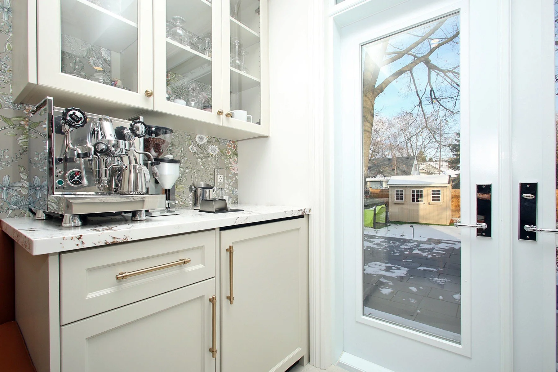 Bright white coffee bar with brass pulls, quartz counter, floral backsplash, and glazed door to the yard.