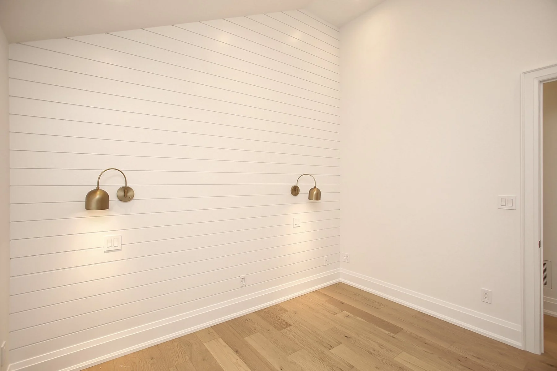 Bedroom corner highlighting a white shiplap wall with two brushed brass sconces above oak hardwood flooring.
