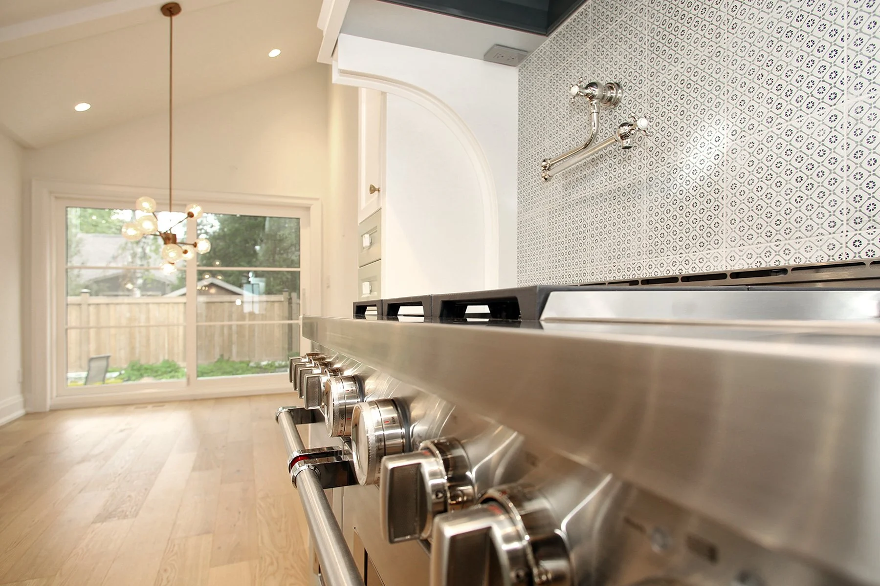 Close perspective along a stainless pro range toward a dining nook lit by a brass globe chandelier in a vaulted space.
