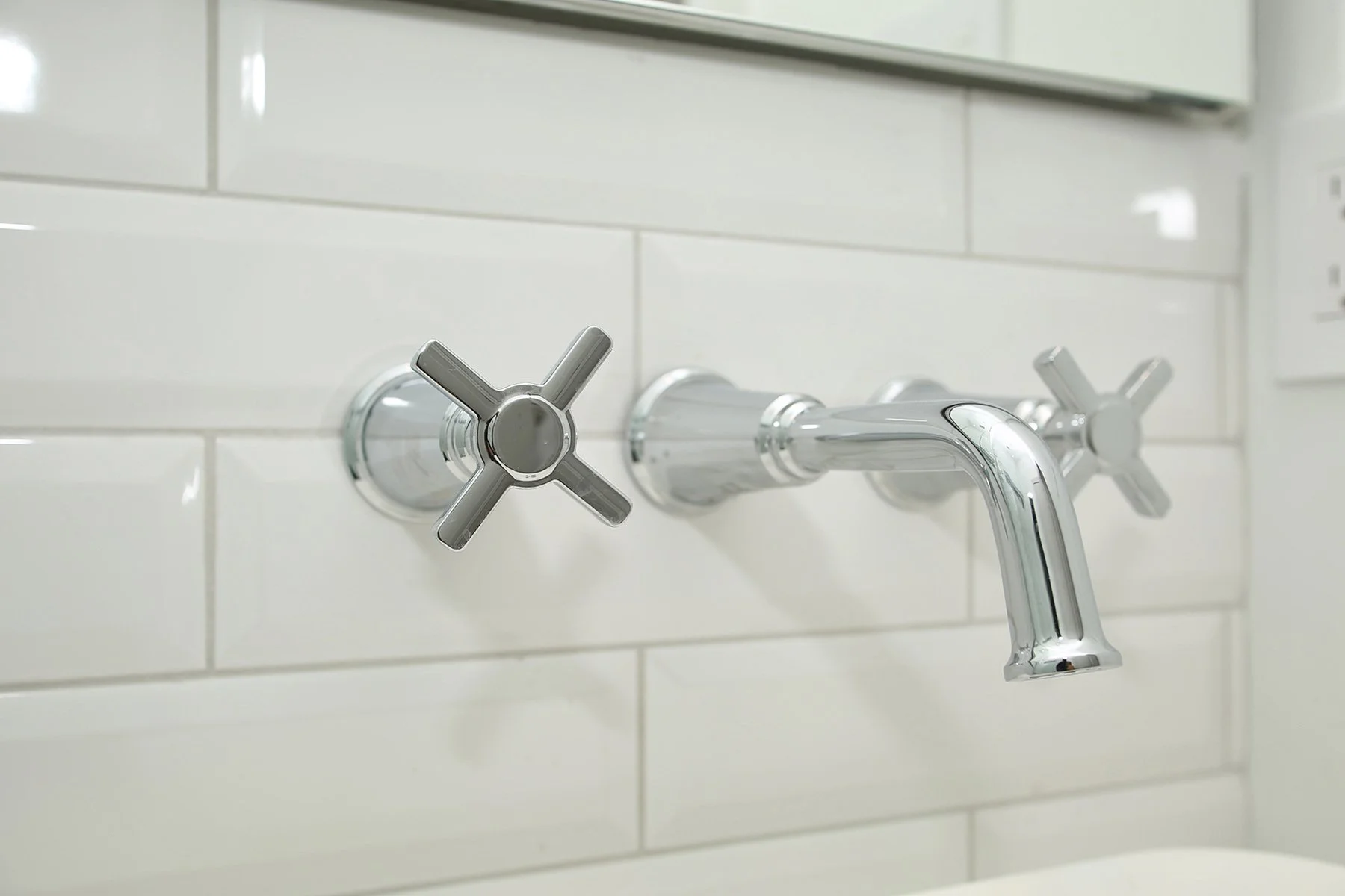 Close up of a polished chrome wall mount bathroom faucet with cross handles on white beveled subway tile.