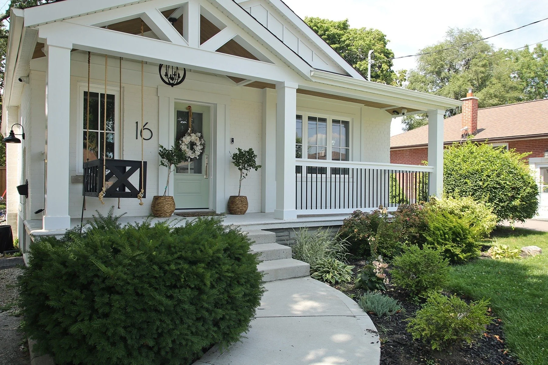 Front porch of the white bungalow with a sage green door, wreath, house number sixteen, and a timber truss gable above.