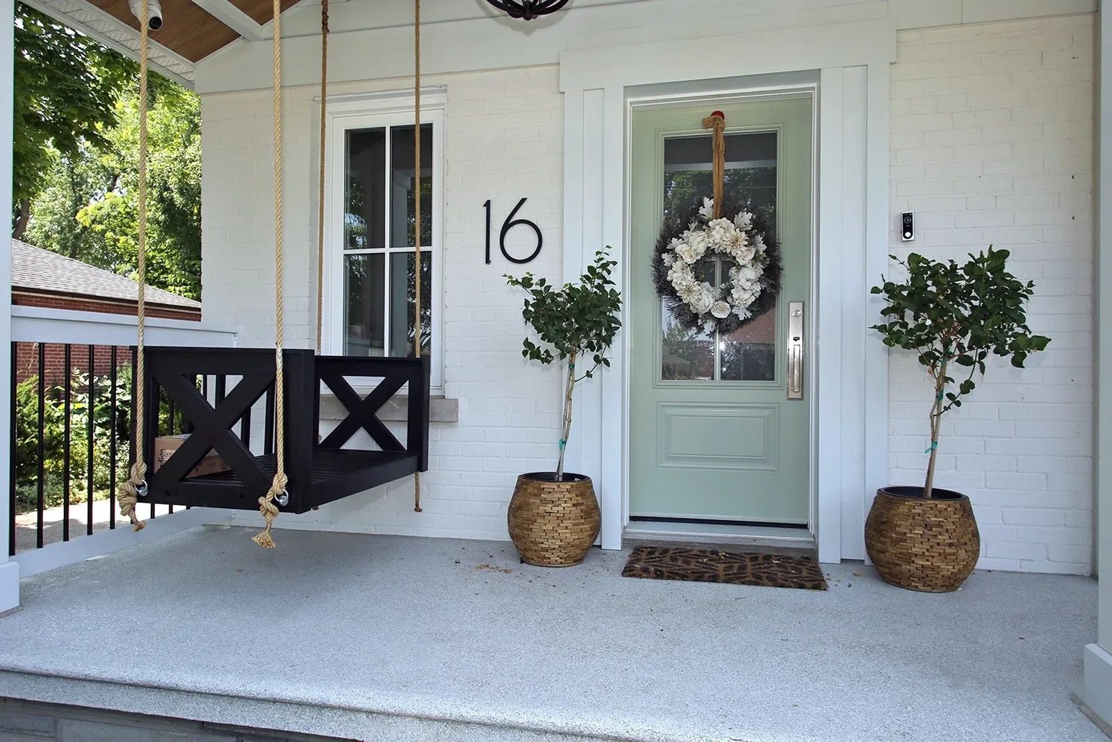 White brick porch with a sage green front door, black wood swing, and symmetrical topiary planters welcoming guests.