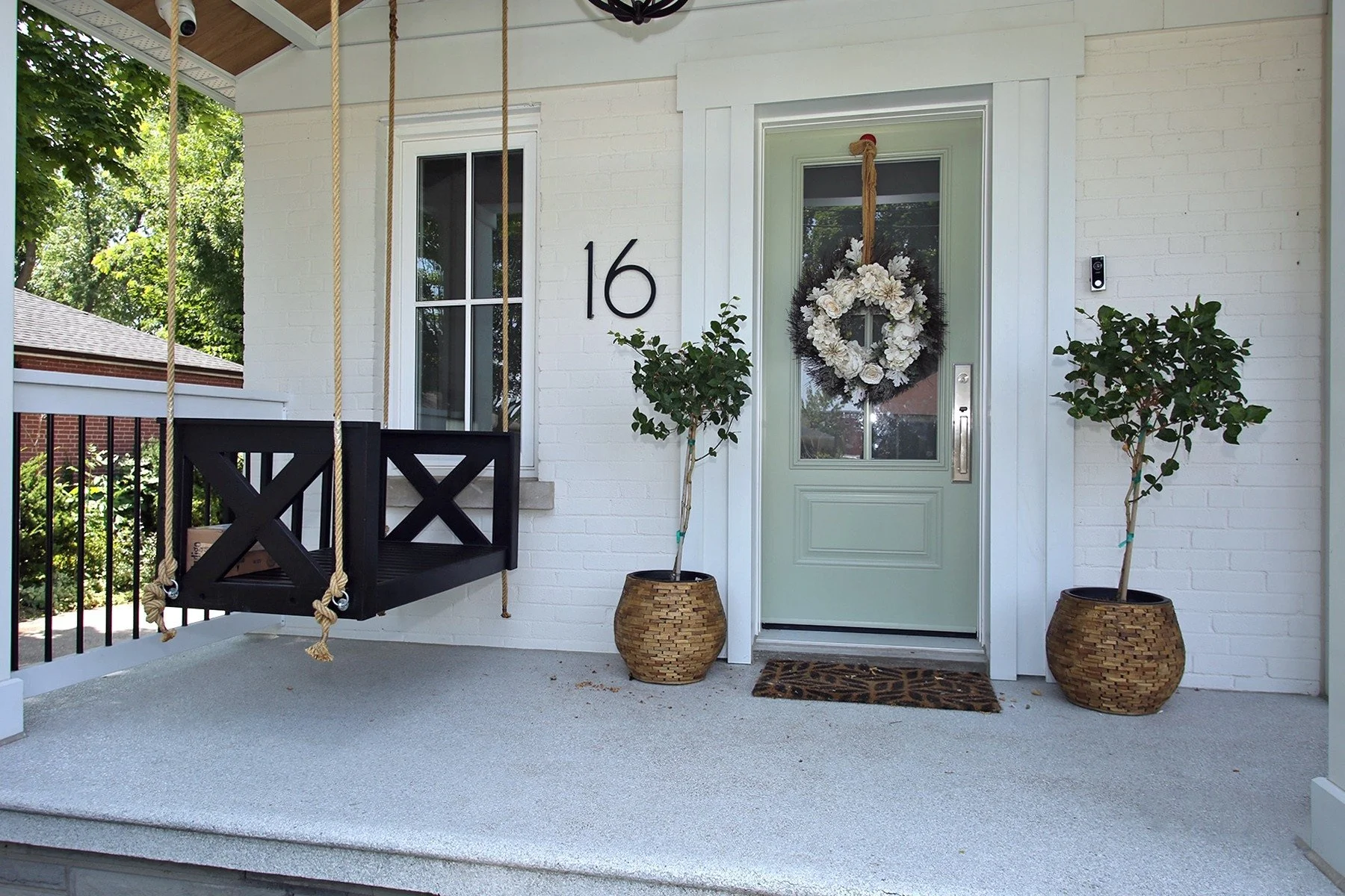 White brick porch with a sage green front door, black wood swing, and symmetrical topiary planters welcoming guests.
