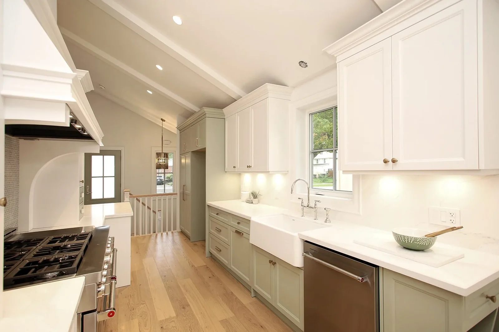 Bright galley kitchen with sage green lower cabinets, a white apron sink at the window, and veined quartz counters.