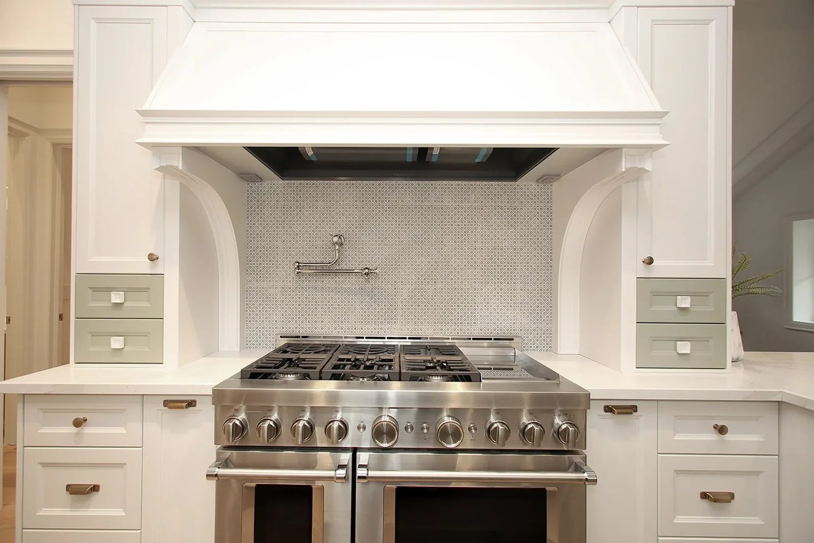 Symmetrical white range wall centres a stainless pro range under a custom hood with sage accent drawers on either side.