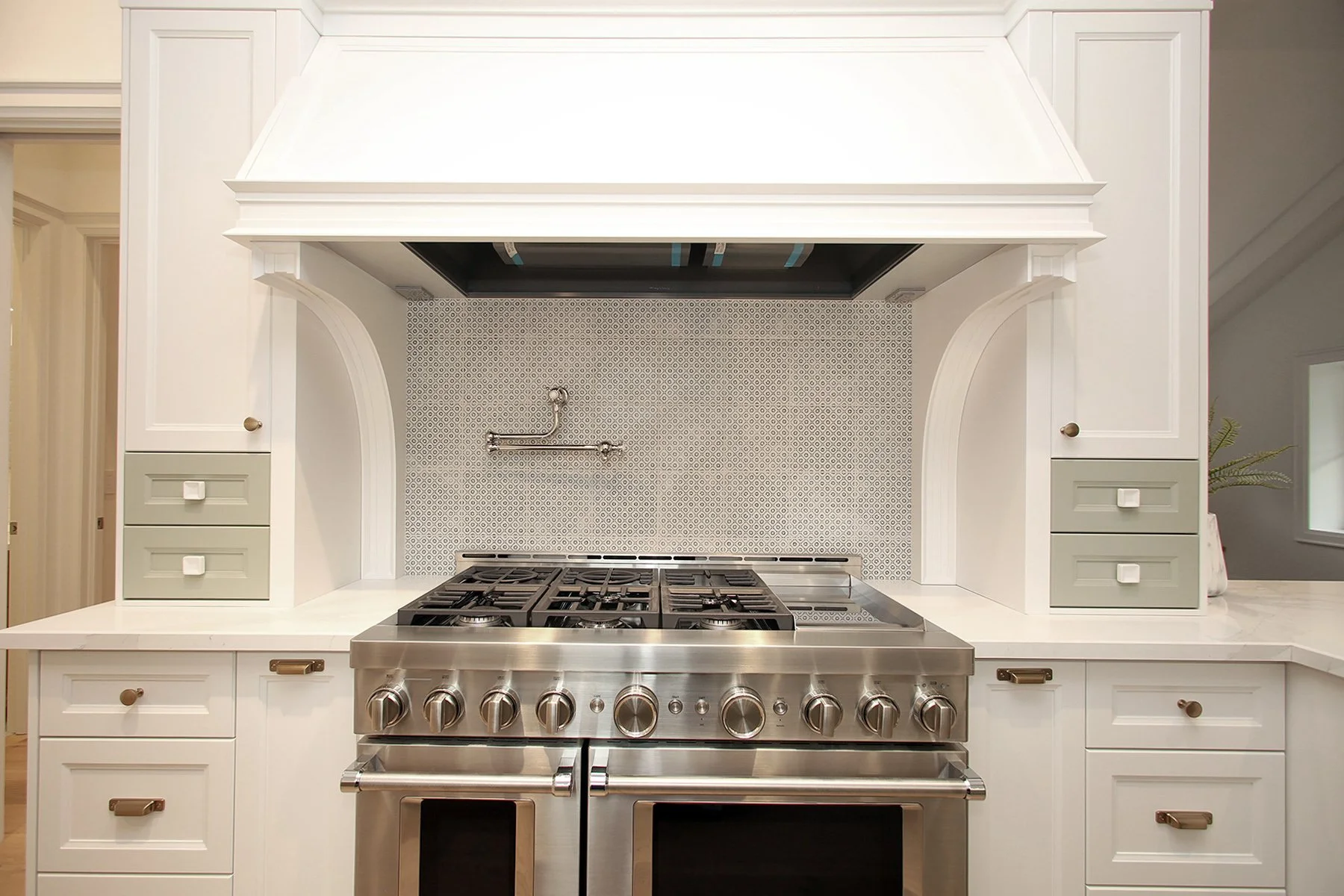 Symmetrical white range wall centres a stainless pro range under a custom hood with sage accent drawers on either side.
