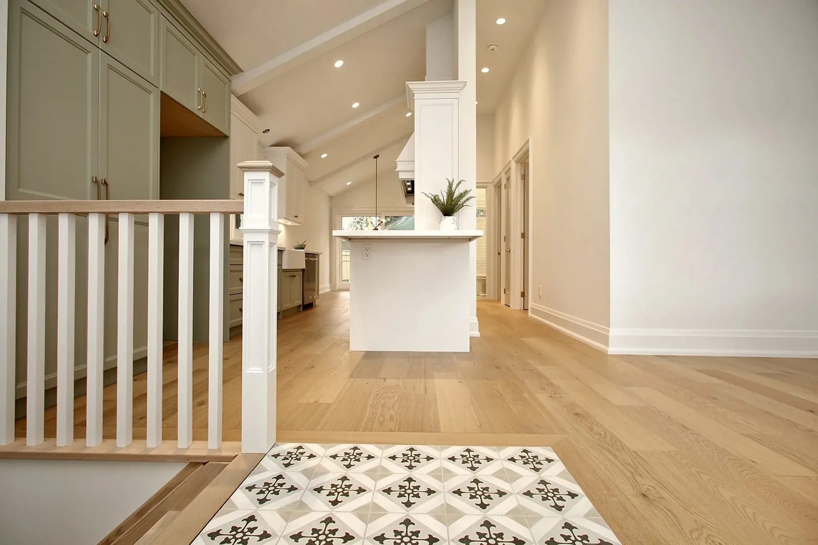 From the stair landing toward the kitchen island, sage pantry wall, oak floors, and patterned tile at the foyer entry.