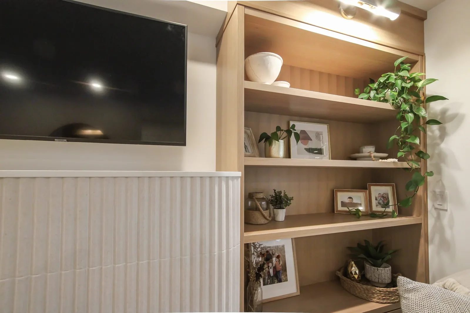 Warm oak shelving and a wall mounted TV beside white fluted panelling in a finished living room.