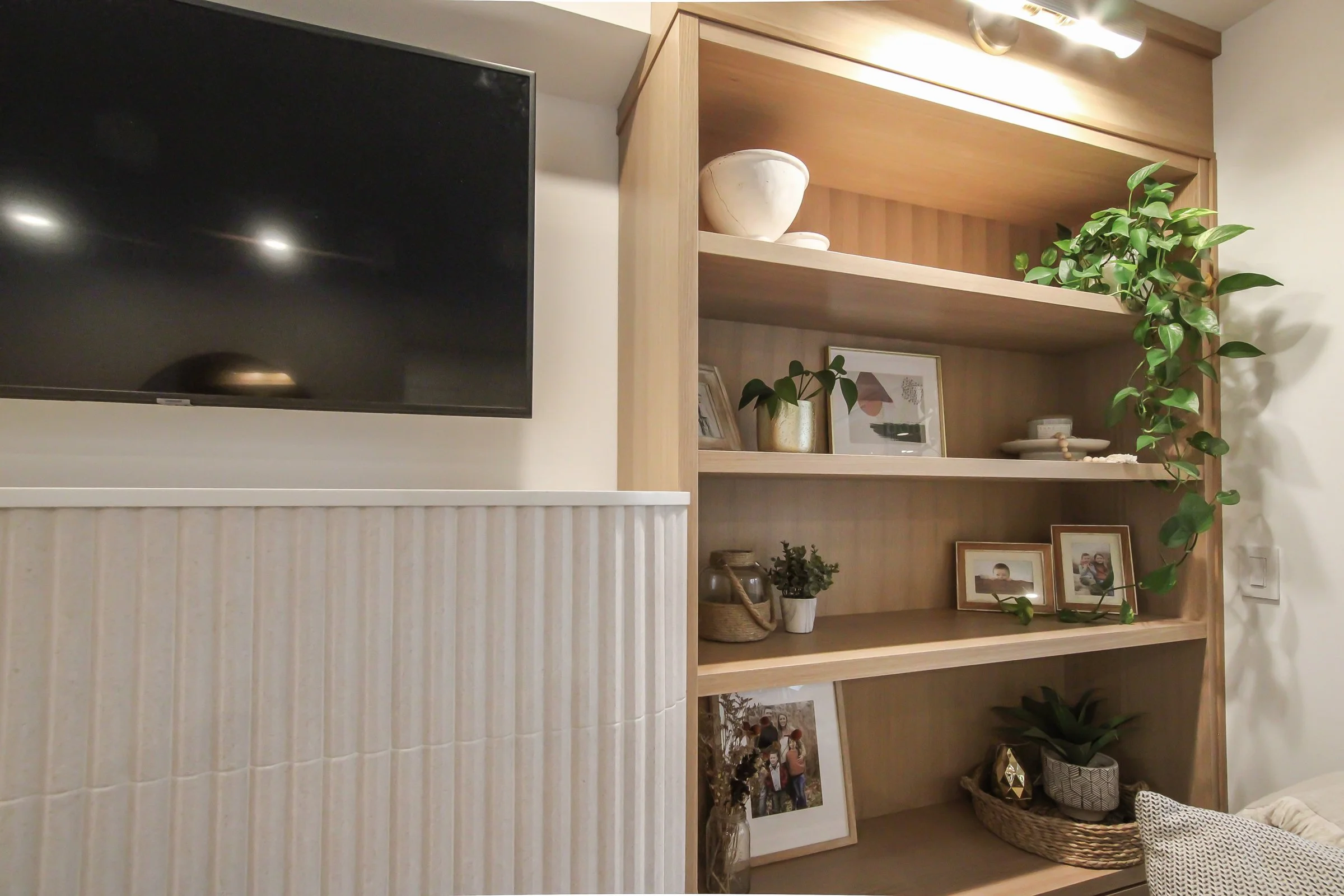 Warm oak shelving and a wall mounted TV beside white fluted panelling in a finished living room.