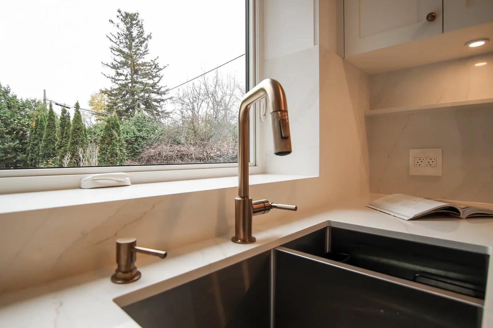 Warm white kitchen sink nook with quartz counters, bronze tap, open shelving, and a large window.