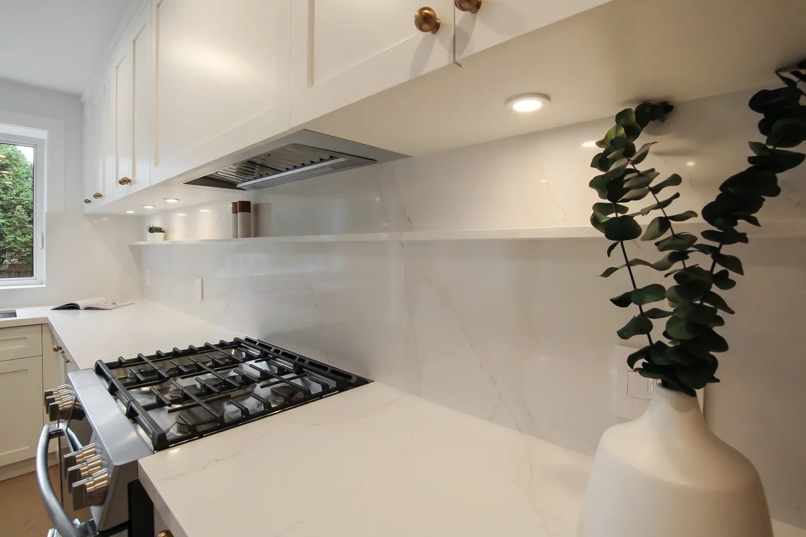 Bright white kitchen detail with quartz backsplash, gas range, brass hardware and staged greenery.