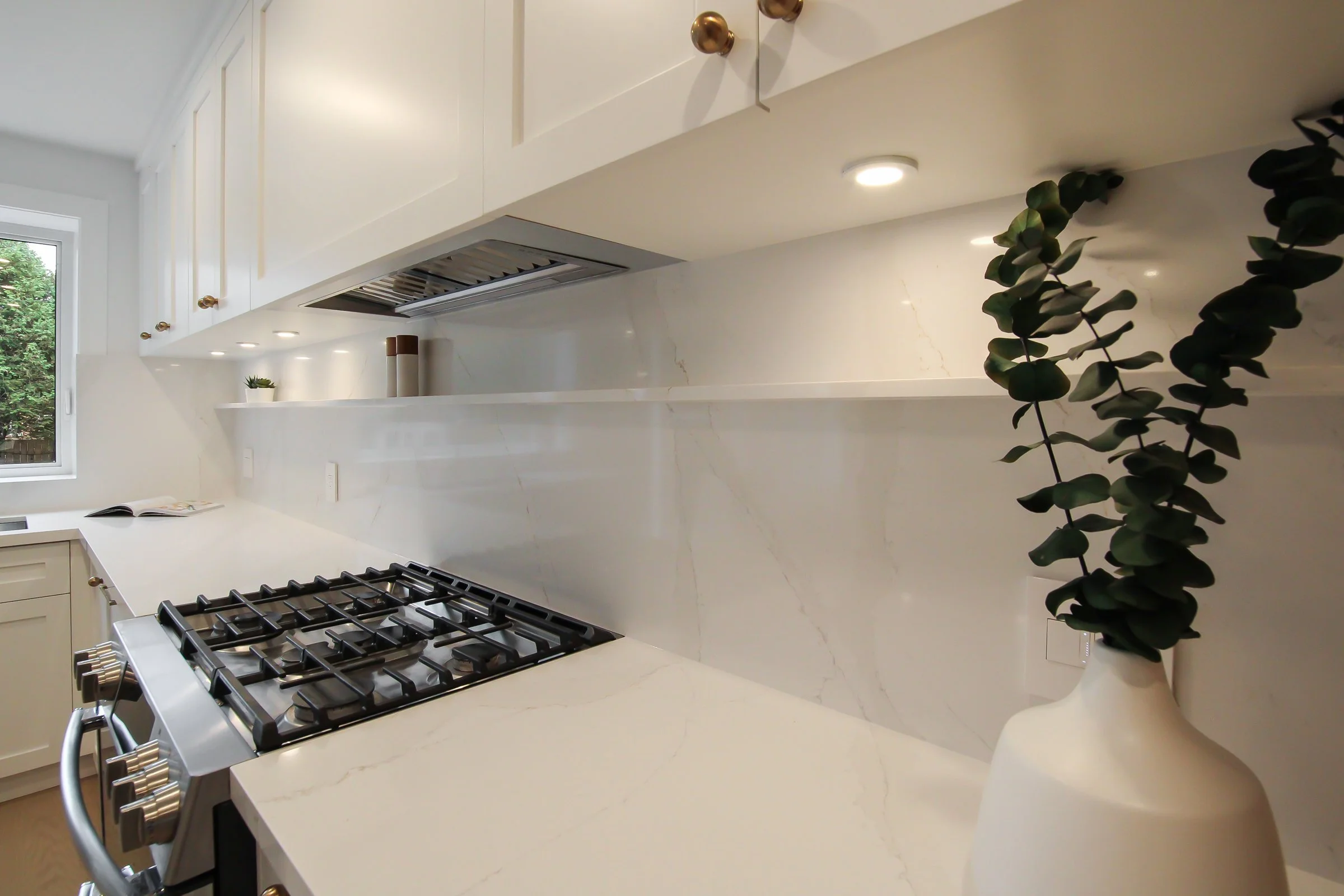 Bright white kitchen detail with quartz backsplash, gas range, brass hardware and staged greenery.