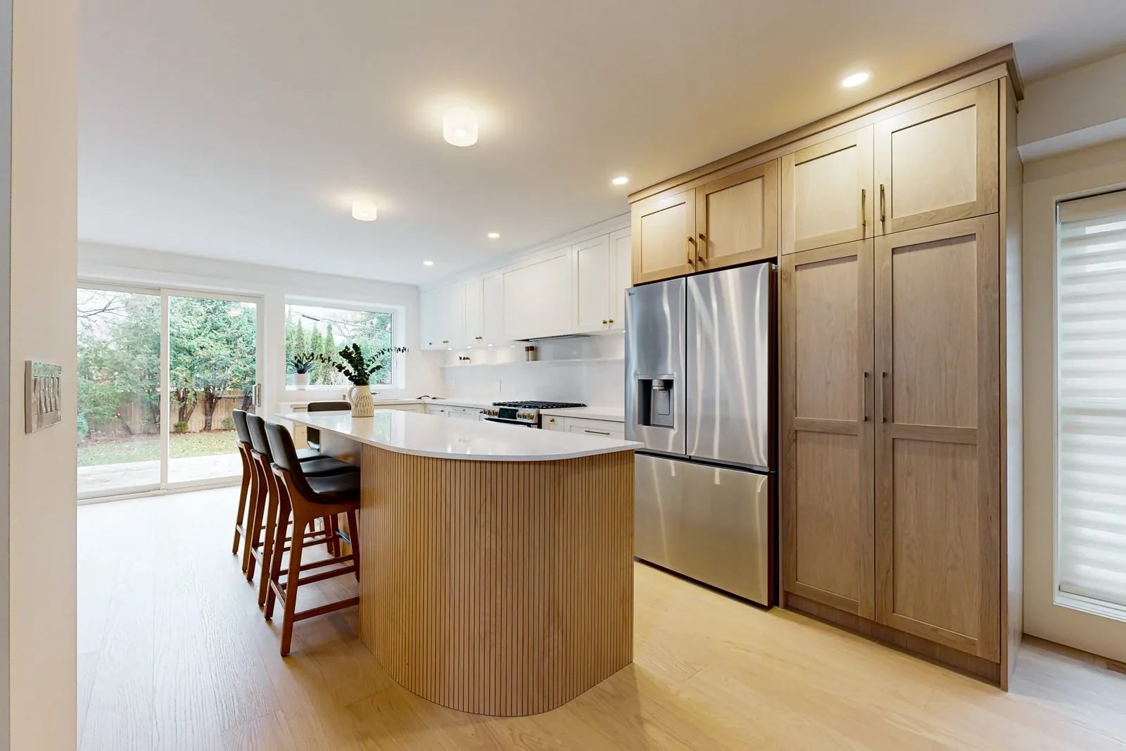 Bright renovated kitchen with white cabinetry, oak pantry wall, curved island and garden views.