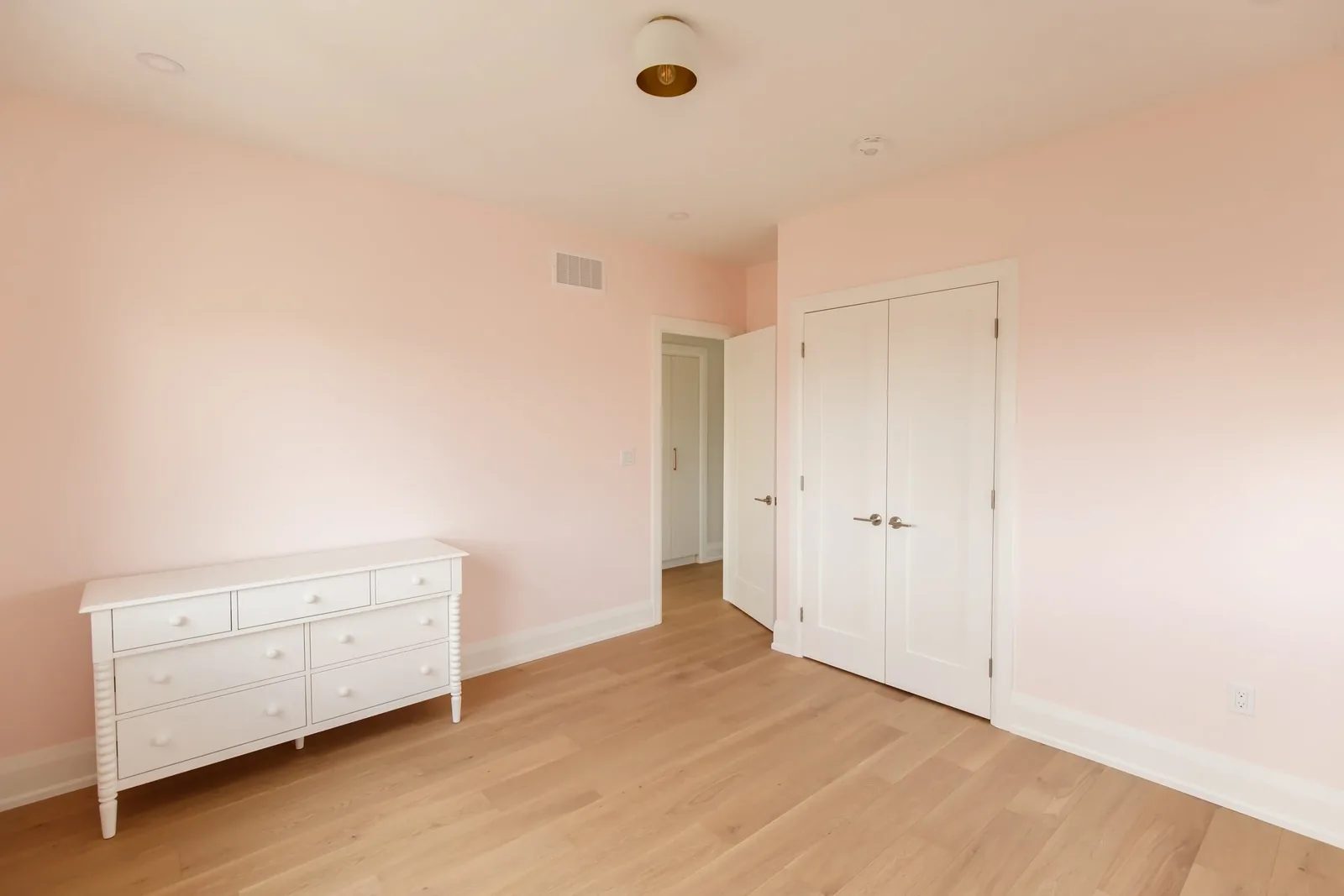 Blush pink bedroom with white spindle dresser, double closet doors and oak floor.