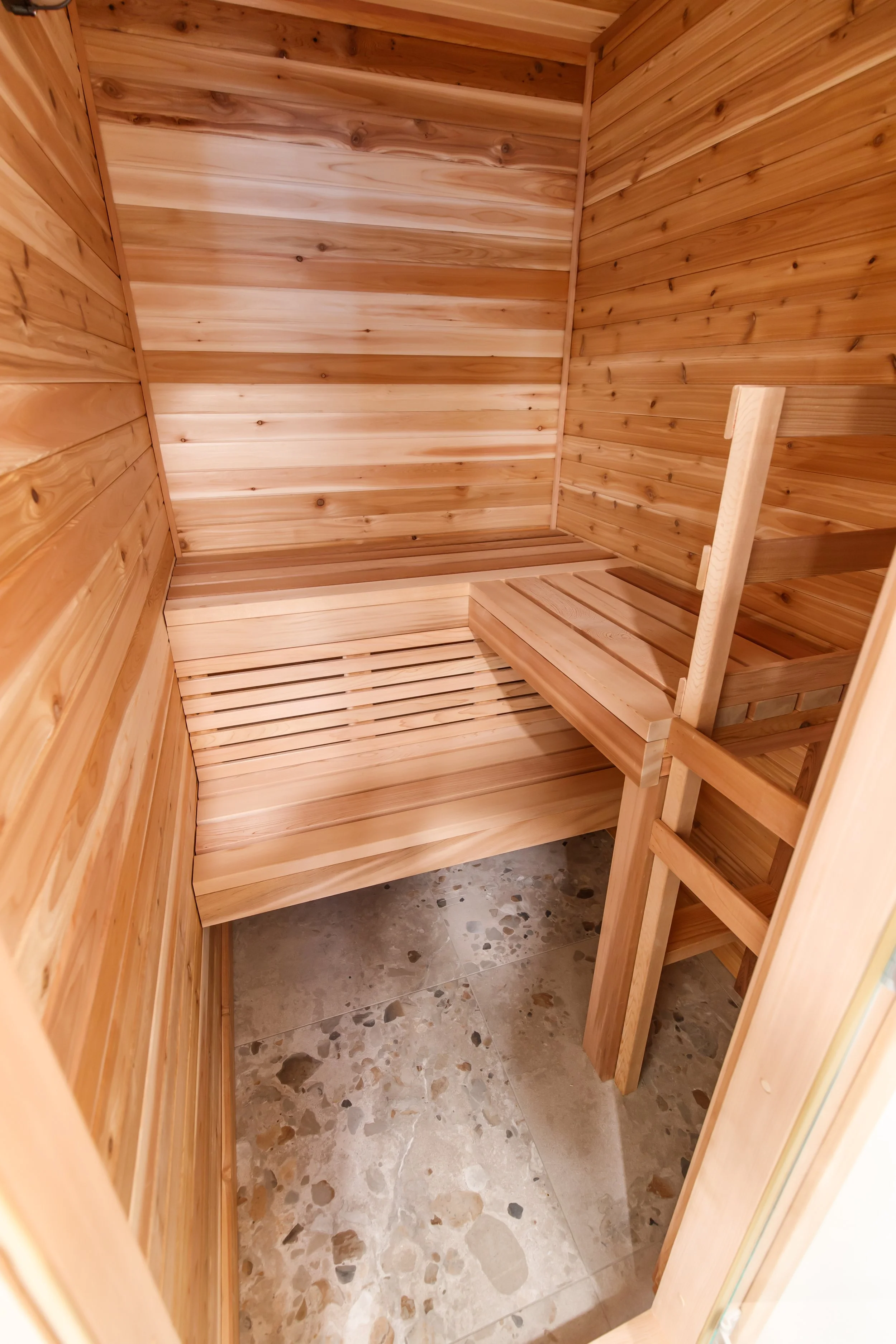 Interior of a cedar home sauna with tiered benches and a vertical cedar ladder over terrazzo tile.