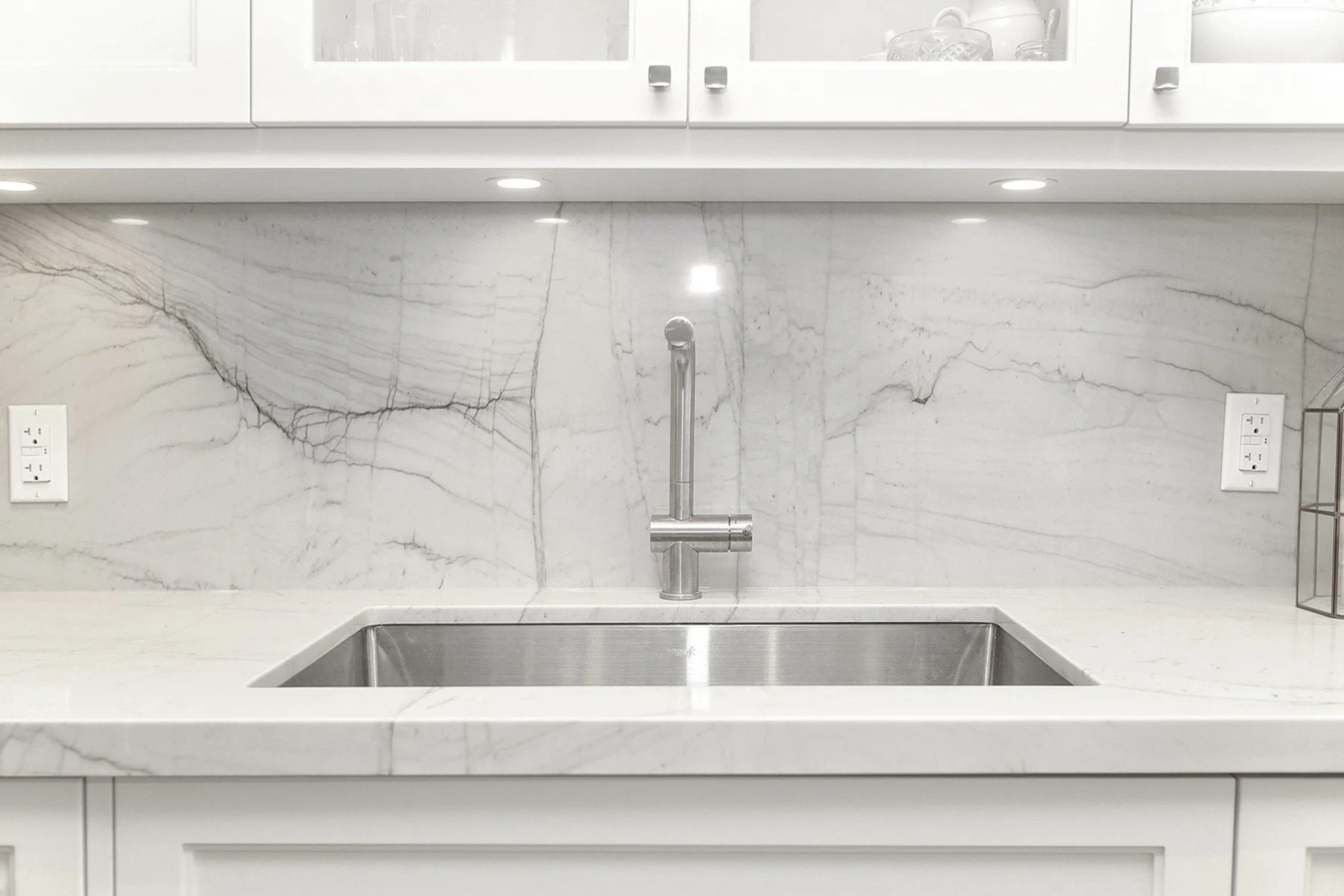 White kitchen sink wall with veined stone backsplash, stainless fixtures, and clean modern millwork.