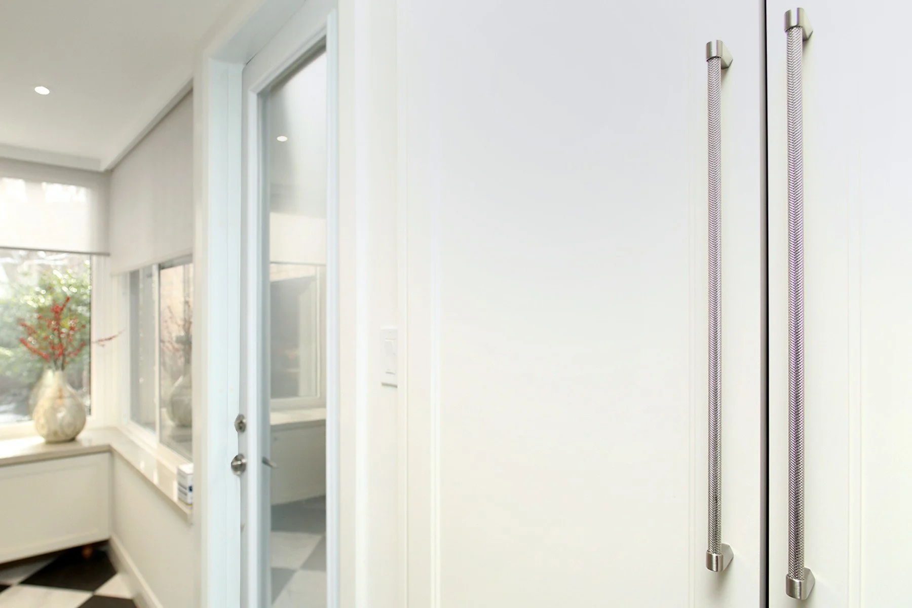 Bright white hallway with a glass door, tall brushed metal pulls, and checkerboard tile by the windows.