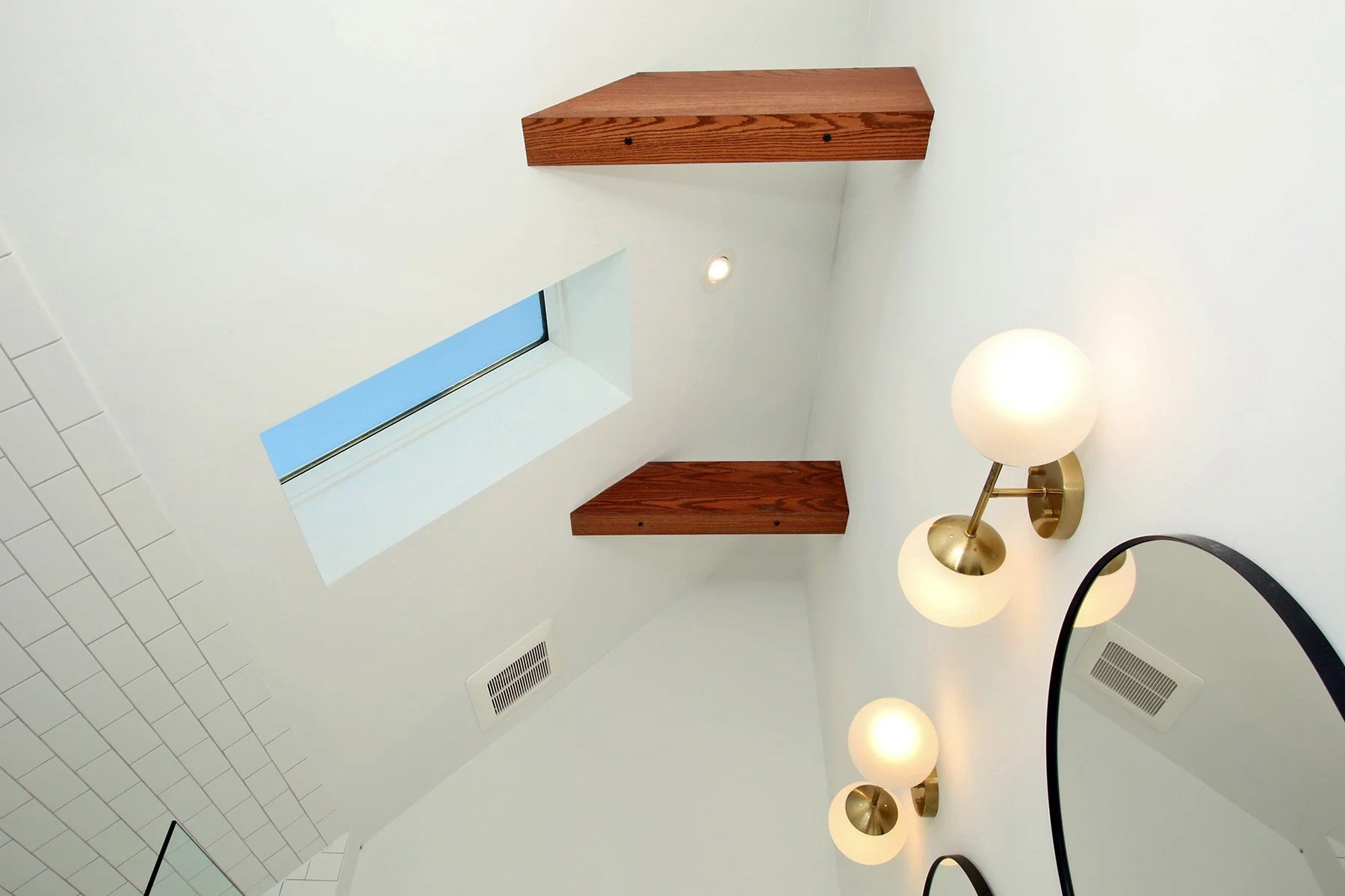 Warm white bathroom detail with a skylight, wood shelves, brass sconces, and a black mirror.