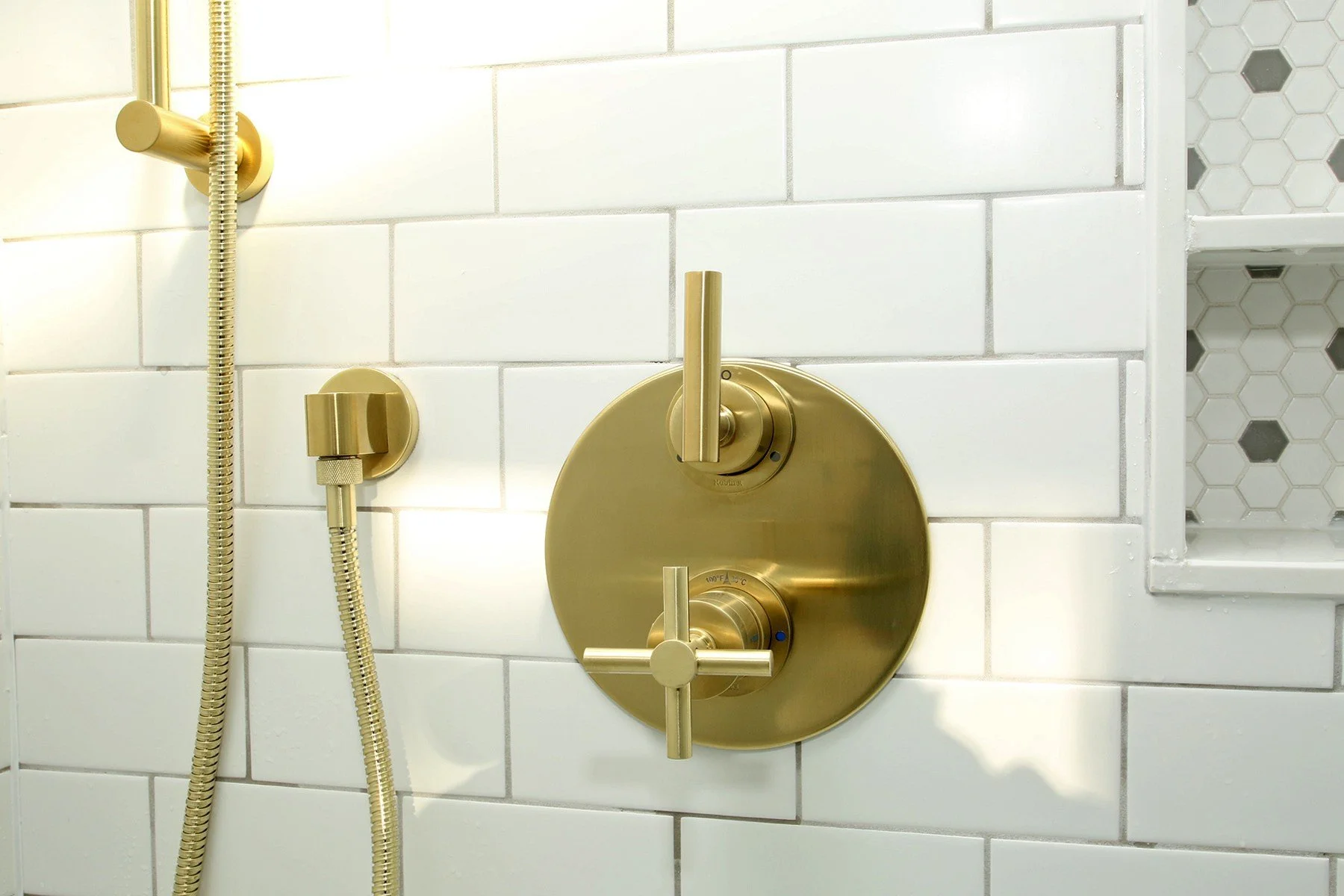Brushed brass shower controls set against warm white subway tile with a tiled niche in the corner.