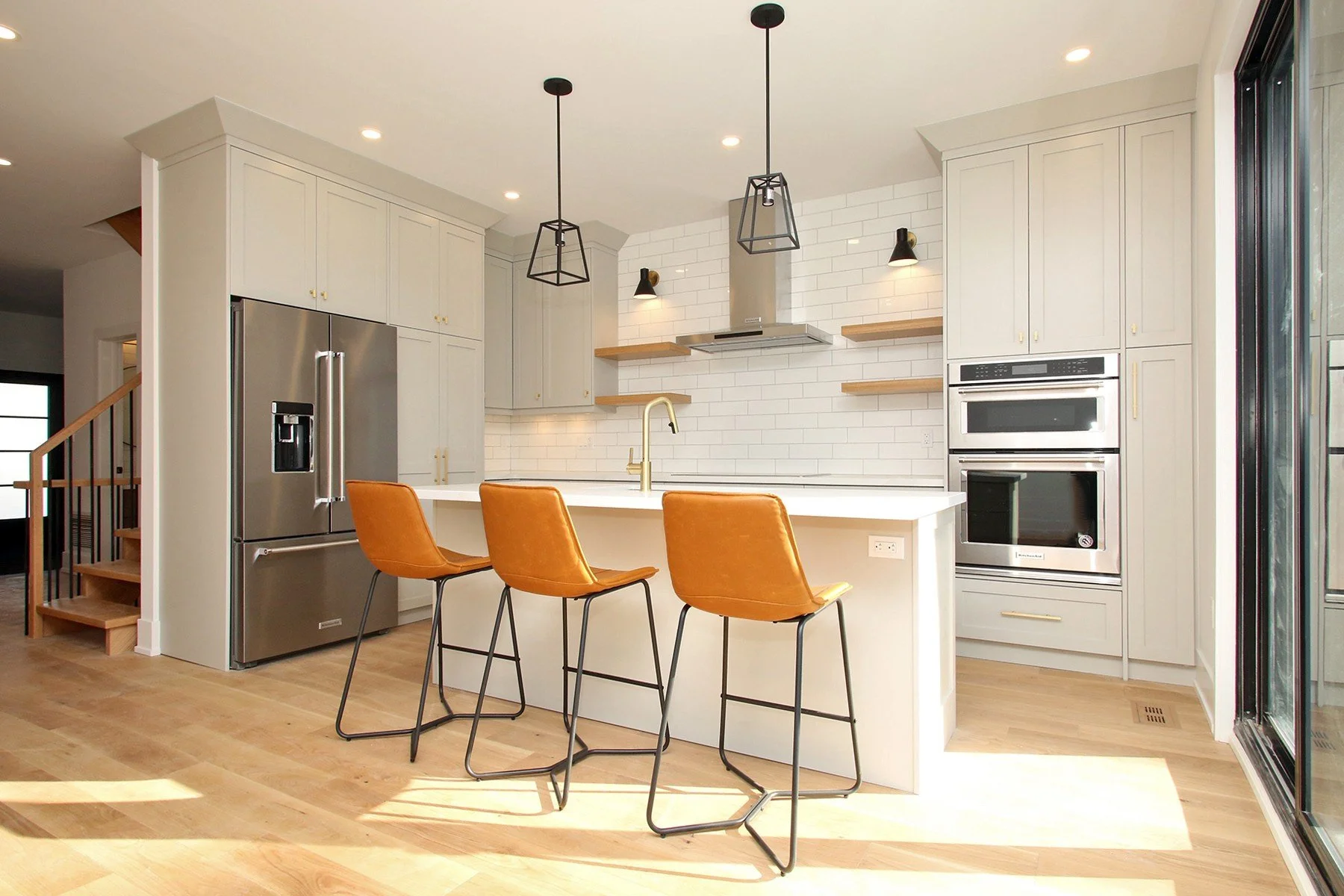 Finished warm white kitchen with oak flooring, brass accents, an island and three tan stools.