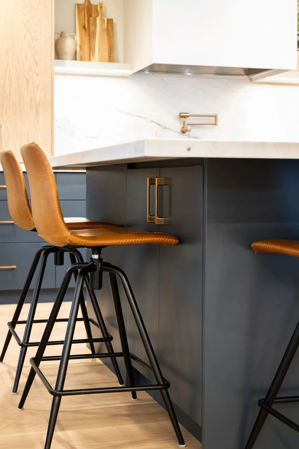 Amber leather counter stools tucked under the mitred stone island edge, with navy cabinetry and a brass pull behind.