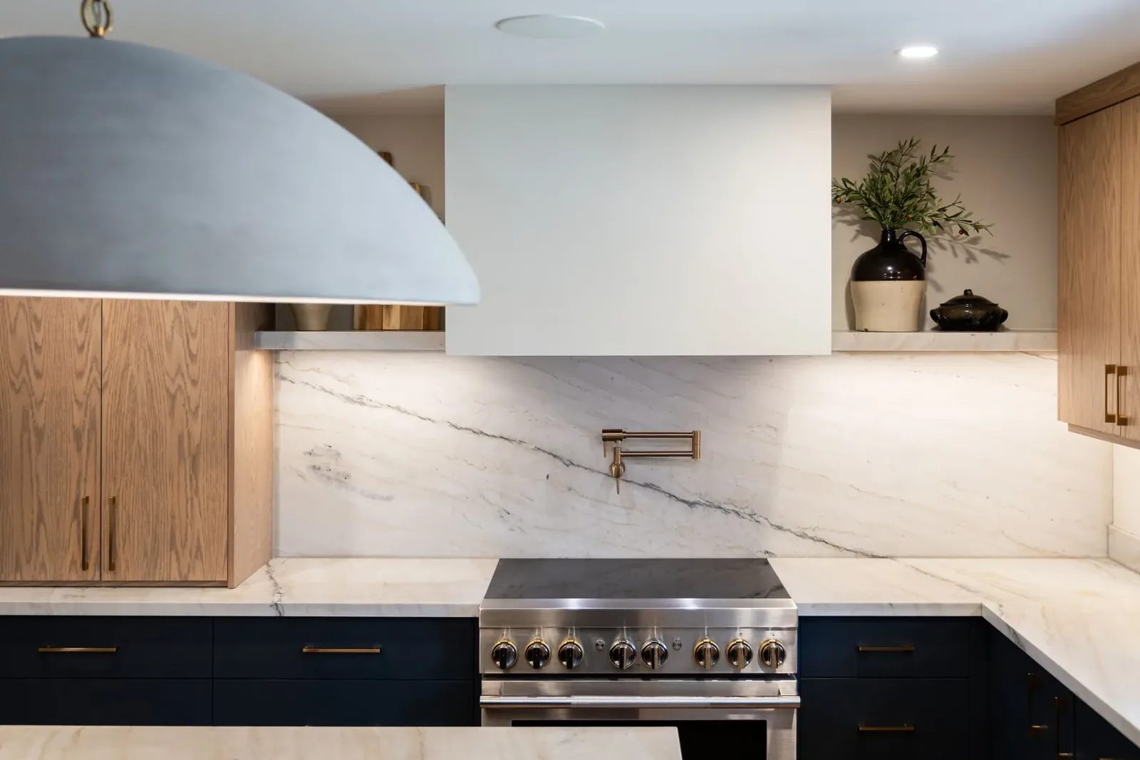 Range wall in full, with a white plaster hood, a bookmatched stone slab backsplash, a brass pot filler, white oak cabinetry and a stainless pro range.