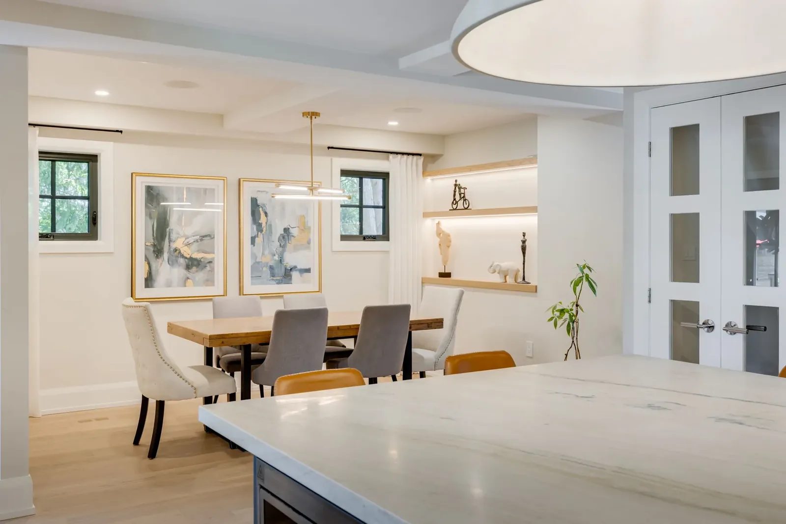 Dining room seen past the kitchen island, with a live-edge wood table, upholstered chairs, framed art and a lit white oak display niche beyond.