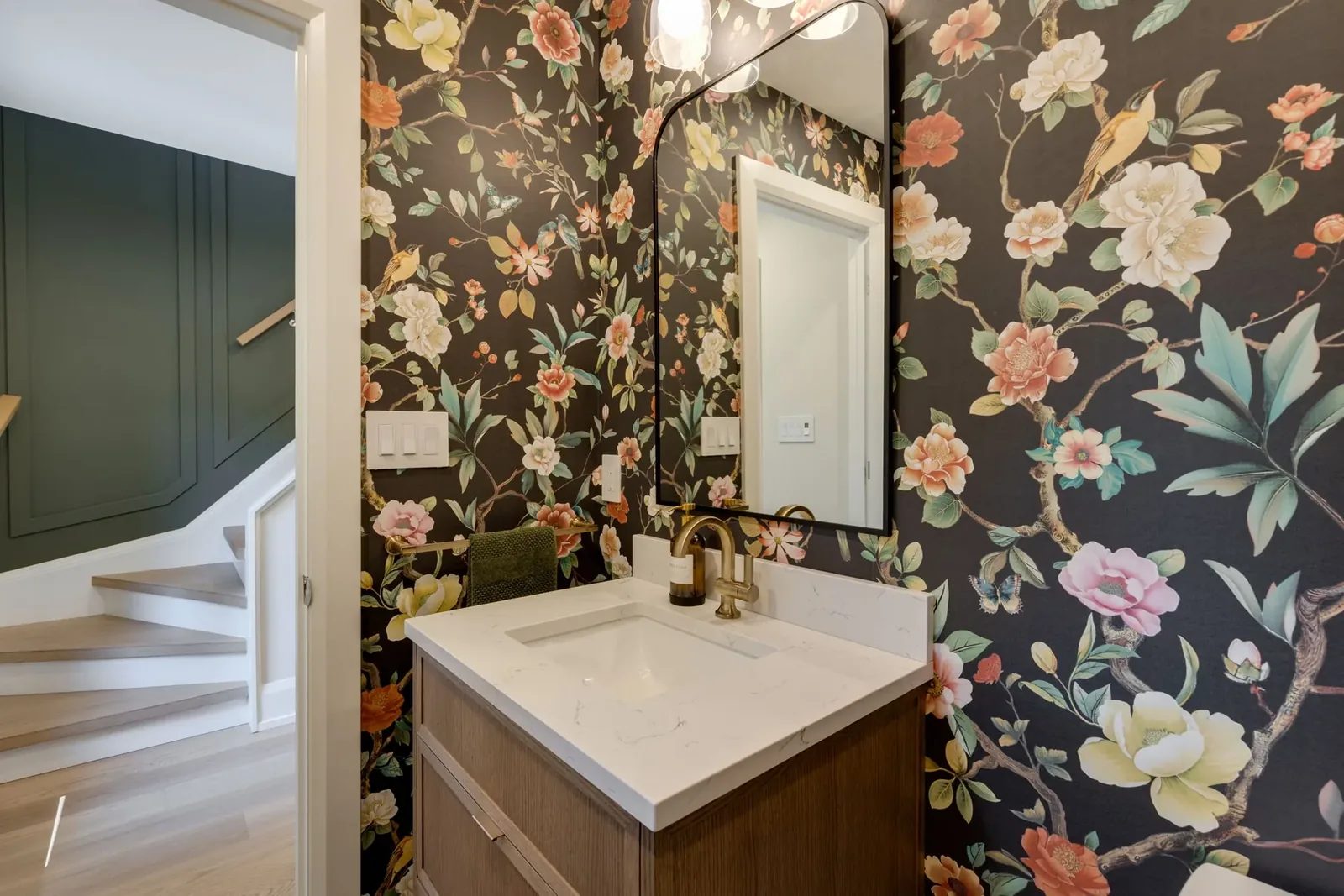 Powder room seen from the stair side, so the deep green panelled staircase and the floral-papered walnut vanity read in one frame.