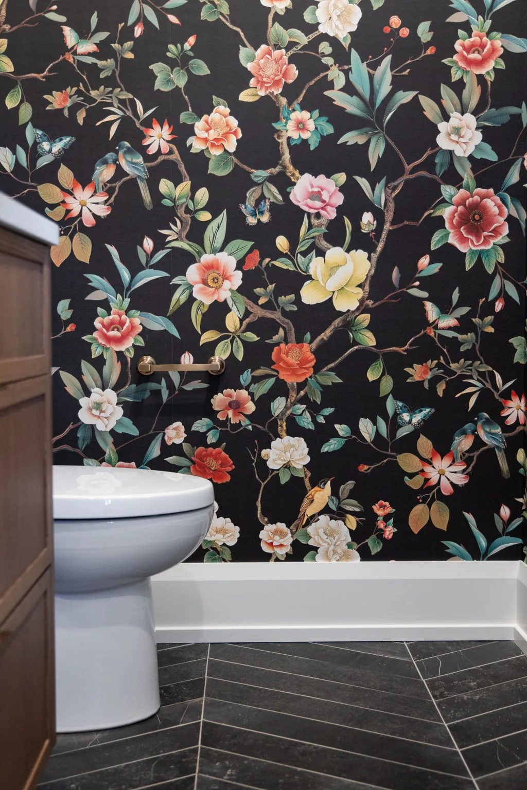 Powder room with a white toilet set against the dark floral wallpaper of birds and blossoms, over a charcoal herringbone tile floor.