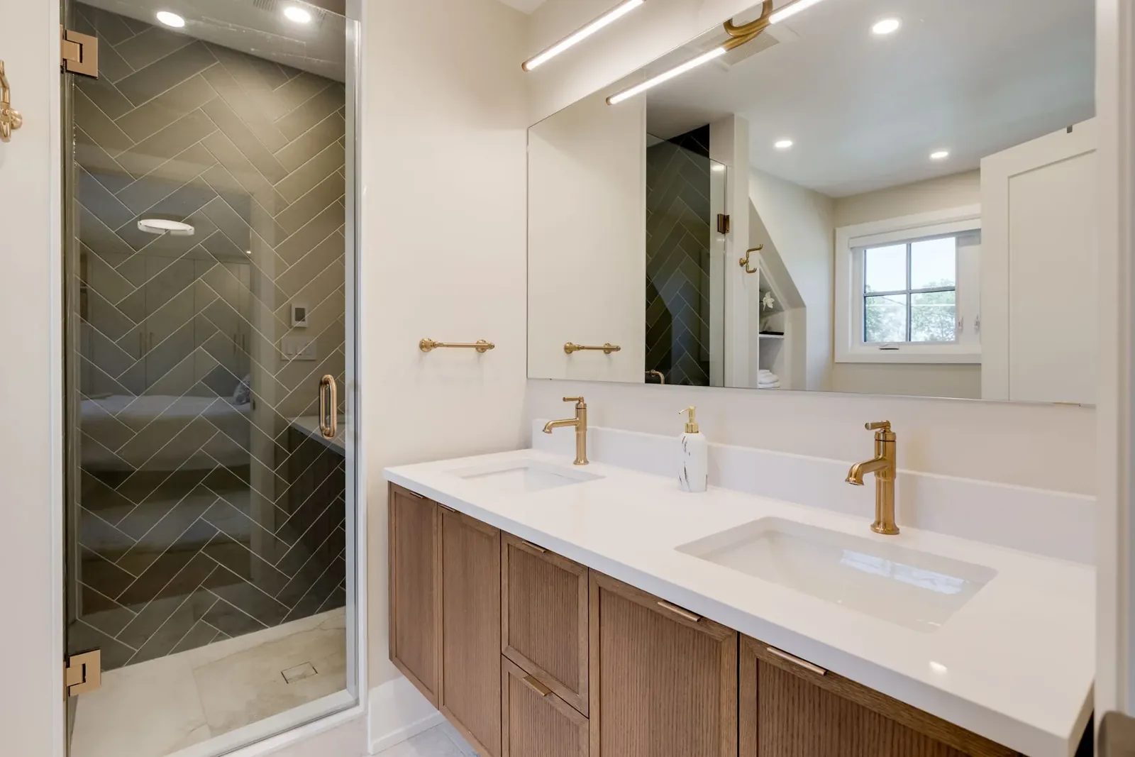 Bathroom with a reeded walnut double vanity, a stone counter, brass tapware and a frameless glass shower clad in green herringbone tile.