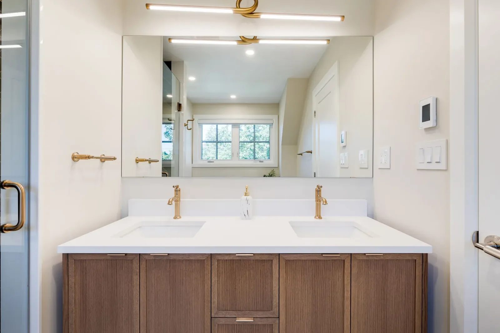 Reeded walnut double vanity straight on, with twin brass faucets, a stone counter and a full-width mirror under a linear brass light.