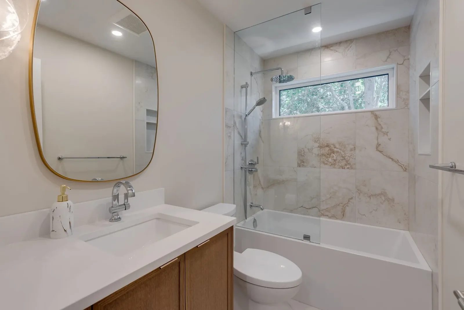Bathroom with a single oak vanity, an oval brass mirror and a tub set into a stone-tiled alcove behind a glass panel, under a transom window.