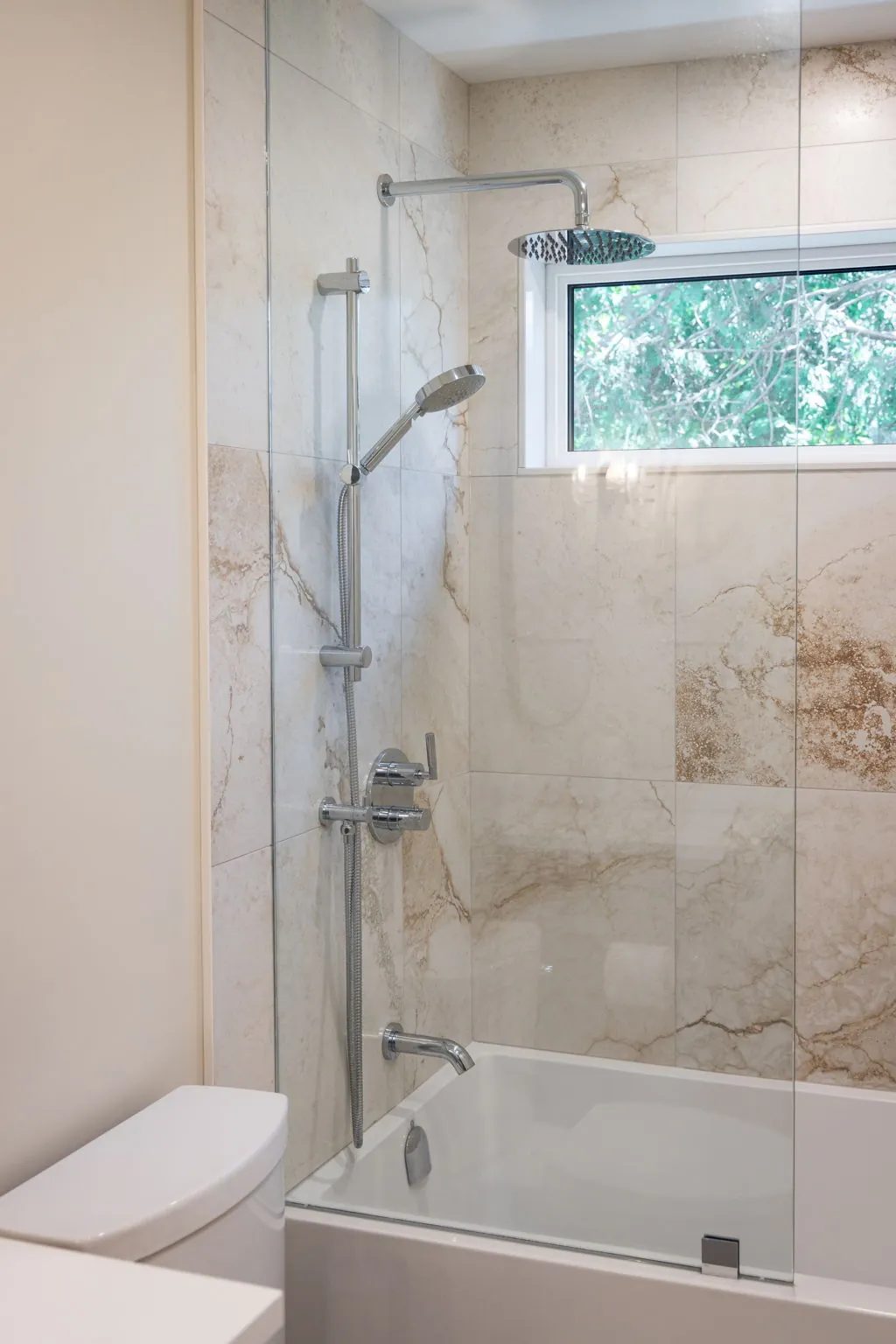 Tub and shower alcove lined in large-format stone-look tile, with a chrome rainfall head, a hand shower on a riser and a glass screen.