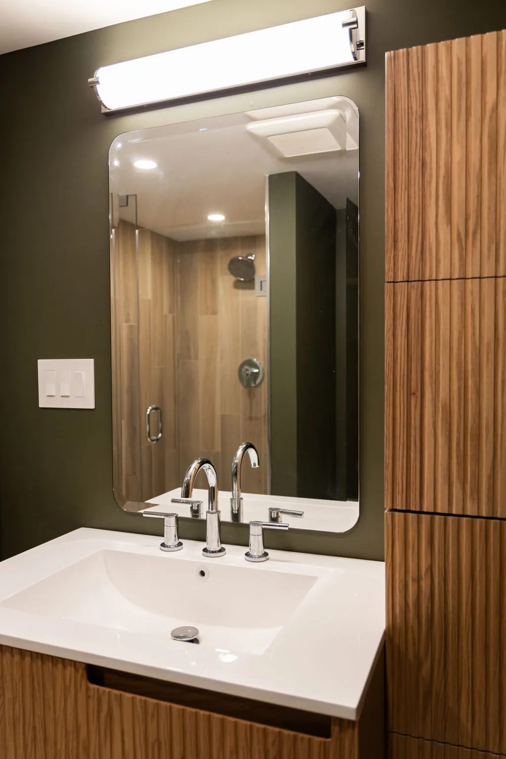 Ensuite vanity with a chrome faucet and a frameless mirror reflecting the wood-look tile shower, beside a reeded wood tall cabinet.