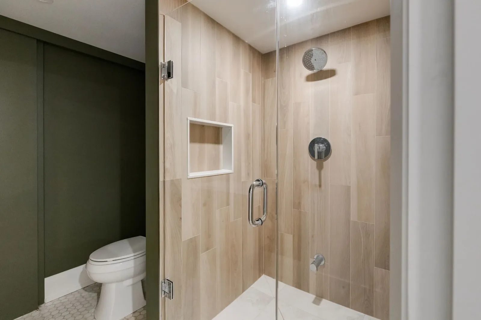 Wood-look tile shower behind frameless glass, with a chrome rainfall head, a round valve and a recessed niche, the green wall beyond.