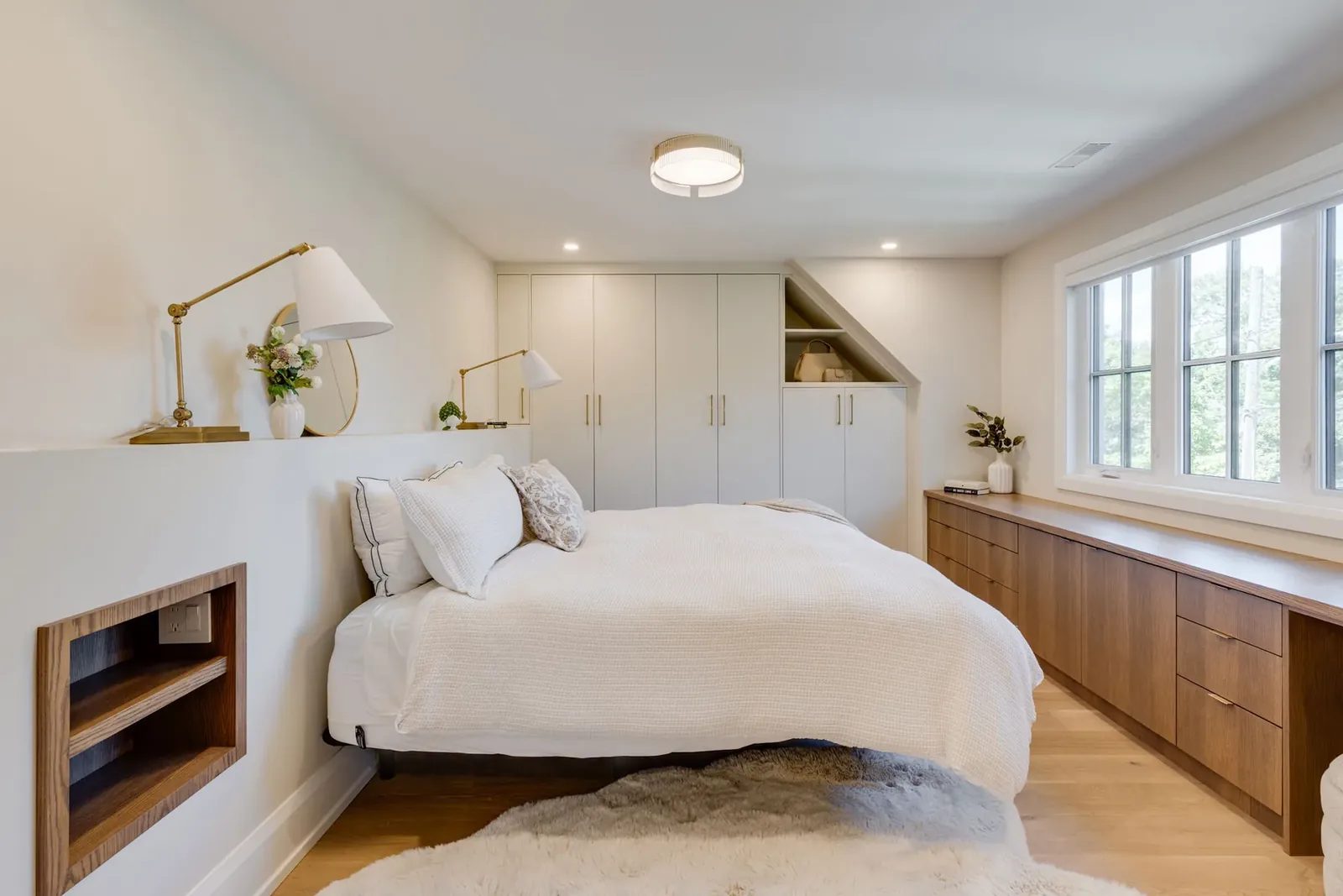 Bedroom with a wall of white built-in wardrobes, a walnut dresser run under a wide window and a built-in oak niche in the low headboard wall.