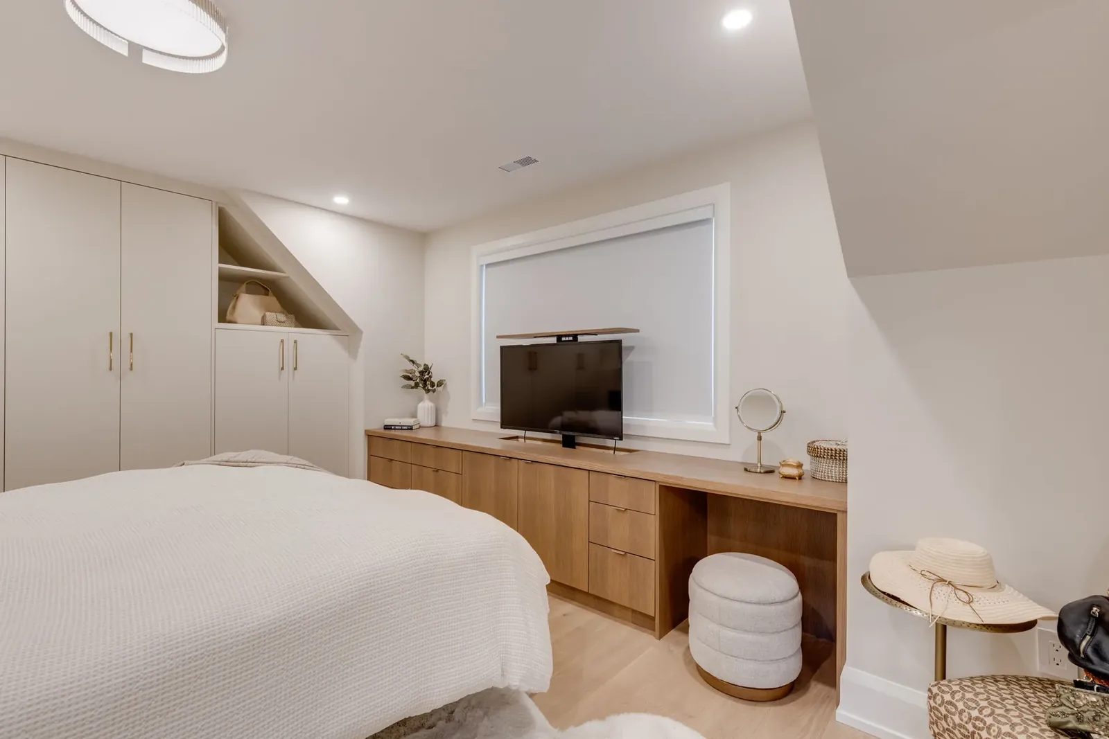 Bedroom with a low white oak console holding a lift-up television, a knee-hole desk with an upholstered stool and tall built-in wardrobes.