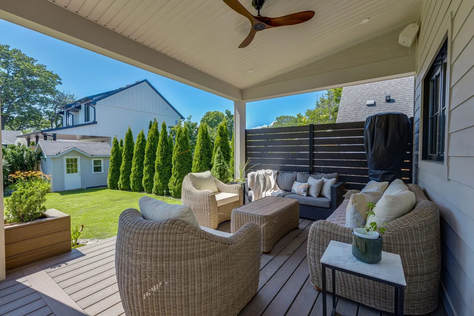 Covered rear porch with woven lounge seating, a dark-bladed ceiling fan, a horizontal slat privacy screen and a view over the lawn.