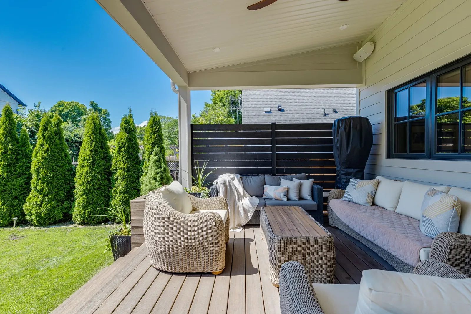 Covered porch from the garden side, its composite deck holding woven sofas and chairs beneath the painted soffit and slat screen.