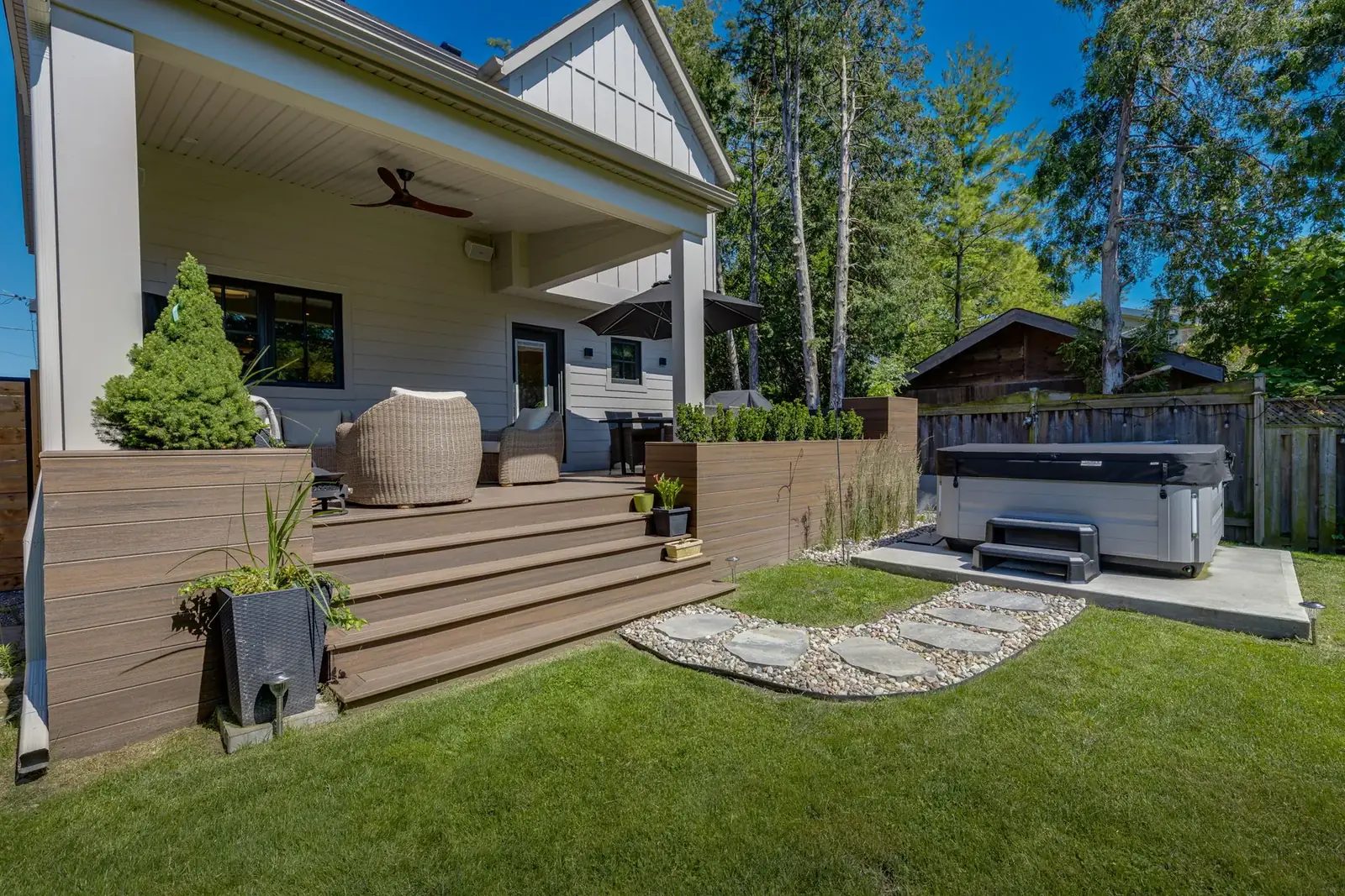 Backyard seen from the lawn, with the covered porch and raised composite deck, planted borders, a stone path and a hot tub on its pad.